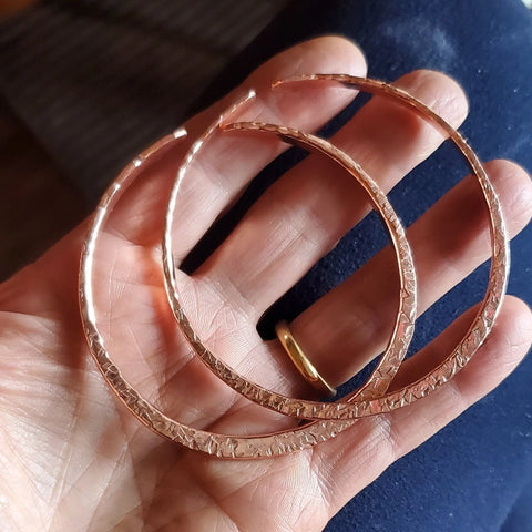 A hand displays two large, textured copper hoop earrings. The wearer also sports a simple gold ring and an Alexa Martha Designs Stackable Hammered Double Canted Angle Bangle, set against a softly blurred background.