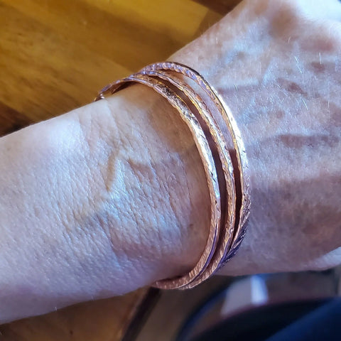 A close-up shows a wrist adorned with three Stackable Hammered Double Canted Angle Bangles by Alexa Martha Designs, set against a wooden background.