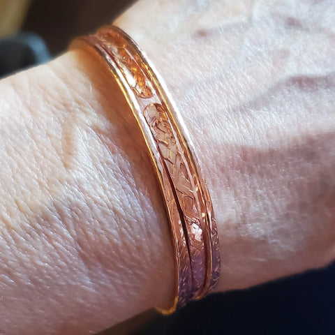 A close-up shows an older adults wrist with two thin, intricately patterned Stackable Hammered Double Canted Angle Bangles by Alexa Martha Designs. Wrinkled skin and veins highlight the artisan copper bracelets elegant style.