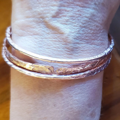 A wrist displays three Stackable Hammered Double Canted Angle Bangles by Alexa Martha Designs in rose gold, their subtle patterns and elegant texture standing out against a softly blurred background.