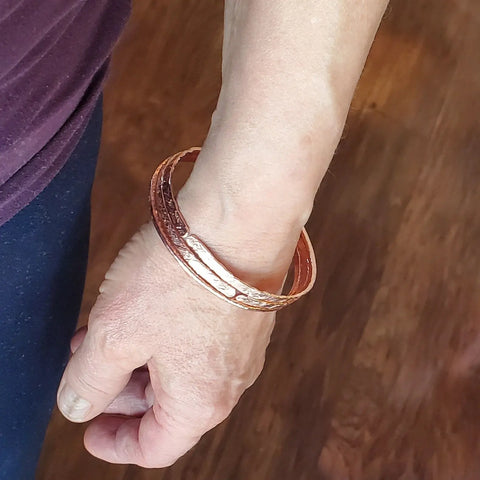 A left hand and wrist model the Alexa Martha Designs Stackable Hammered Double Canted Angle Bangles against a wooden floor, with the person wearing a purple shirt and dark pants.