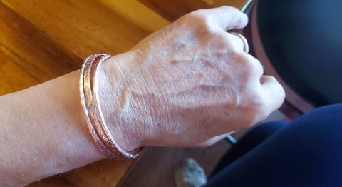 A close-up of a hand resting on wood, wearing Alexa Martha Designs Stackable Hammered Double Canted Angle Bangles in a copper hue.