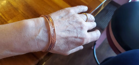 A hand wearing Alexa Martha Designs Stackable Hammered Double Canted Angle Bangles and a ring rests on a wooden table, with part of a chair and the edge of the table in the background.