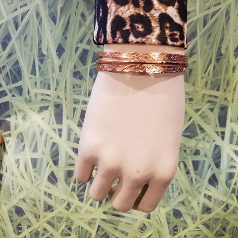 A mannequin hand wears Alexa Martha Designs’ Stackable Hammered Double Canted Angle Bangles and a long-sleeve leopard print top, set against a green grass-patterned background.