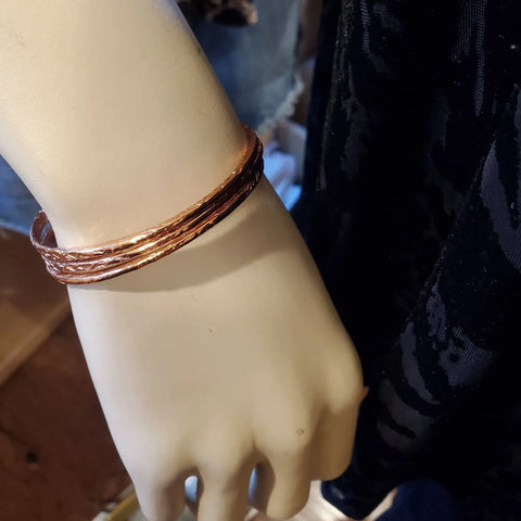 A mannequin hand showcases Stackable Hammered Double Canted Angle Bangles by Alexa Martha Designs in shiny copper, displayed next to a black textured garment.