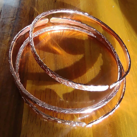 Three Stackable Hammered Double Canted Angle Bangles by Alexa Martha Designs are elegantly stacked on a wooden surface, their shiny finish reflecting sunlight and casting distinct shadows.
