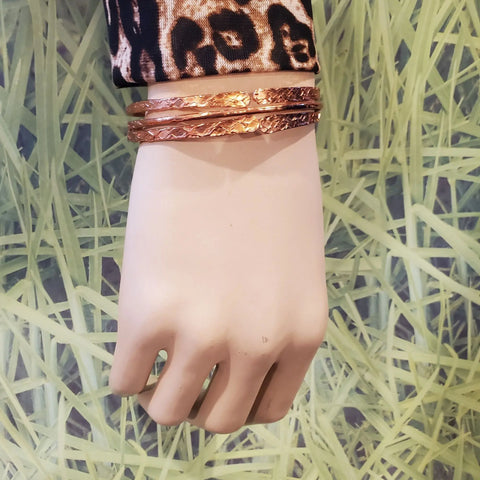 A mannequin hand displays three Stackable Hammered Double Canted Angle Bangles by Alexa Martha Designs, paired with a leopard-print sleeve and set against a green grass-patterned background.