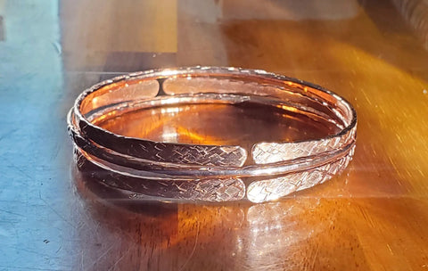 Alexa Martha Designs Stackable Hammered Double Canted Angle Bangles, featuring a textured engraved pattern, are displayed on a polished wooden surface reflecting warm sunlight.