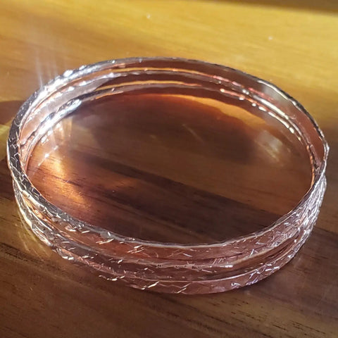 A few Stackable Hammered Double Canted Angle Bangles by Alexa Martha Designs rest on a wooden surface, their shiny finish reflecting sunlight and showcasing artisanal charm.