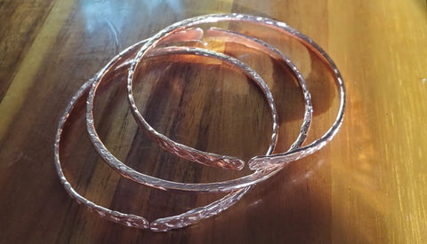 Four Stackable Hammered Double Canted Angle Bangles by Alexa Martha Designs lie loosely overlapped on polished wood, with sunlight casting shimmering reflections and shadows.
