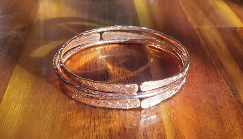 Alexa Martha Designs’ Stackable Hammered Double Canted Angle Bangles, with their textured copper finish, rest on a polished wood surface as gentle sunlight highlights their handmade beauty.