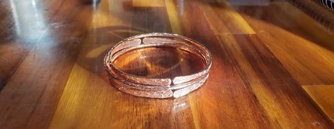 Three Stackable Hammered Double Canted Angle Bangles by Alexa Martha Designs, with a copper finish and textured patterns, are stacked on a polished wooden surface in warm sunlight.