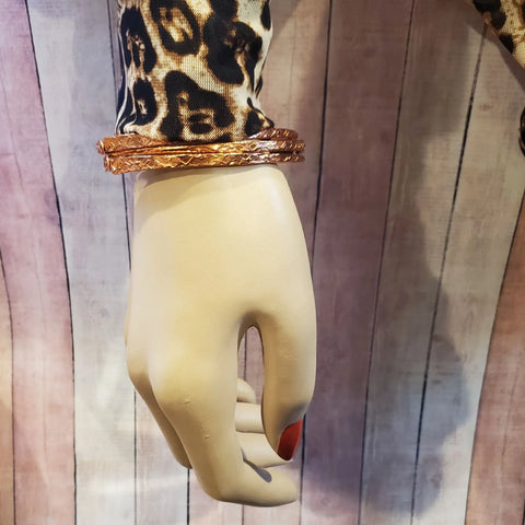 A mannequin hand styled in a leopard print sleeve showcases Alexa Martha Designs’ Stackable Hammered Double Canted Angle Bangles, posed against a wooden plank background.