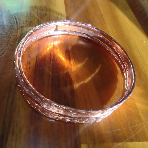 The Stackable Hammered Double Canted Angle Bangles by Alexa Martha Designs, handmade in shiny copper, rest on a polished wooden surface as sunlight casts warm reflections and highlights.