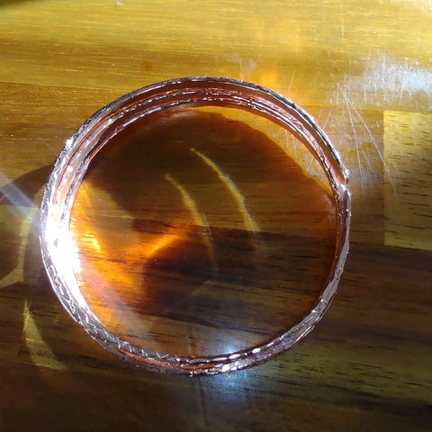 A set of Alexa Martha Designs Stackable Hammered Double Canted Angle Bangles is arranged in a circular stack on a wooden surface, with sunlight highlighting their shiny, textured finish through bright reflections and shadows.