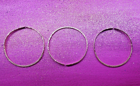 Three metallic bangles from Alexa Martha Designs—Stackable Hammered Double Canted Angle Bangles—are arranged in a row on a purple glittered surface, evoking the elegance of their unique stackable style.
