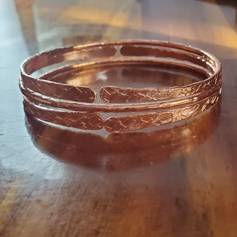 Two shiny Stackable Hammered Double Canted Angle Bangles by Alexa Martha Designs rest side by side on a polished wooden surface, their textured finish reflecting warm light for an elegant, charming touch to any look.