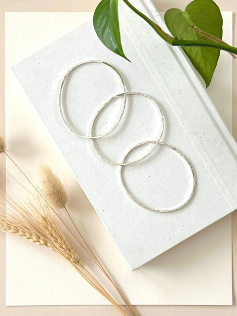 Three HAMMERED SILVER BANGLE BRACELETS by Sarah Cecelia are linked on a white textured book, accented with a green vine and dried wheat—an elegant display of minimalist jewelry on a light, neutral background.