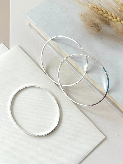 Three Sarah Cecelia HAMMERED SILVER BANGLE BRACELETS are showcased on a textured white surface, complemented by dried grass and stacked books in the background for a minimalist jewelry display.