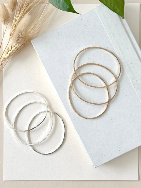 Four HAMMERED SILVER BANGLE BRACELETS by Sarah Cecelia are displayed on white textured paper with dried grass and green leaves, adding a stylish minimalist accent.