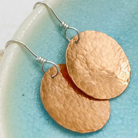 JANECKA Copper Hand Forged Disk Earrings by Janecka feature hand-hammered copper discs and sterling silver ear wires, shown on a turquoise and cream ceramic dish.