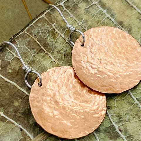 Close-up of Janecka’s JANECKA Copper Hand Forged Disk Earrings, featuring round handmade copper disks and sterling silver ear wires, resting on a green leaf background—showcasing rustic artisan craftsmanship.