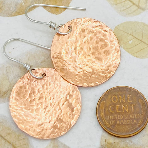 JANECKA Copper Hand Forged Disk Earrings by Janecka, shown beside a U.S. penny for size reference, feature sterling silver ear wires. Displayed on a light, leaf-patterned background—artisan jewelry perfect for any occasion.