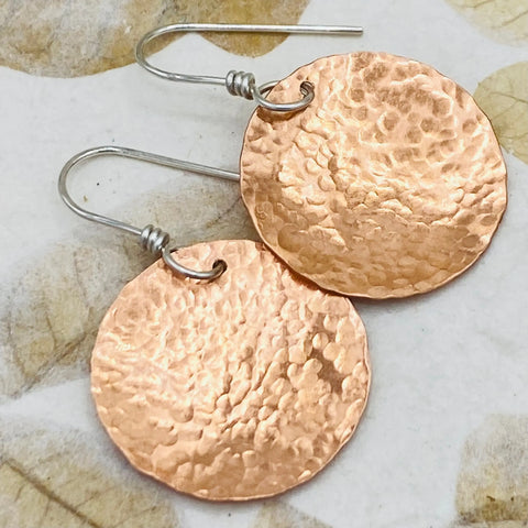 The JANECKA Copper Hand Forged Disk Earrings by Janecka feature round, hammered copper disks with sterling silver ear wires, shown on a light patterned surface.