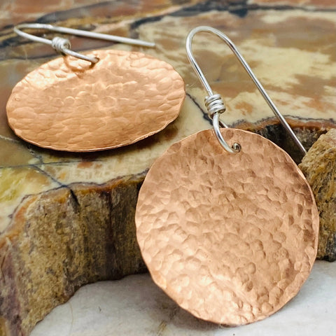 JANECKA Copper Hand Forged Disk Earrings by Janecka feature large, round, hammered copper discs and sterling silver ear wires, shown on textured stone—ideal for fans of artisan crafted jewelry.