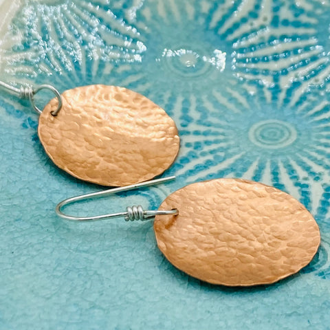 JANECKA Copper Hand Forged Disk Earrings by Janecka, featuring sterling silver ear wires, are displayed on a textured blue ceramic dish with a radiating pattern.
