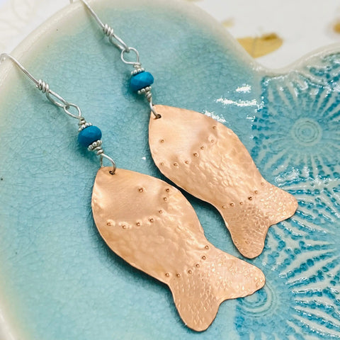 The Janecka JANECKA Copper Hand Forged Fish With Turquoise Earrings feature copper fish and turquoise beads, crafted by hand and shown on a textured turquoise ceramic dish—a unique piece of artisan jewelry.