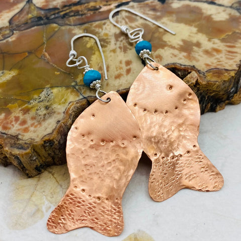The Janecka Copper Hand Forged Fish With Turquoise Earrings, featuring turquoise beads and silver findings, are shown on a polished stone surface.