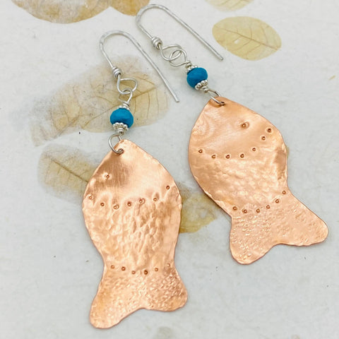 JANECKA Copper Hand Forged Fish With Turquoise Earrings by Janecka feature a hammered finish, dotted accents, and turquoise beads—unique handmade jewelry shown on a leaf-patterned background.
