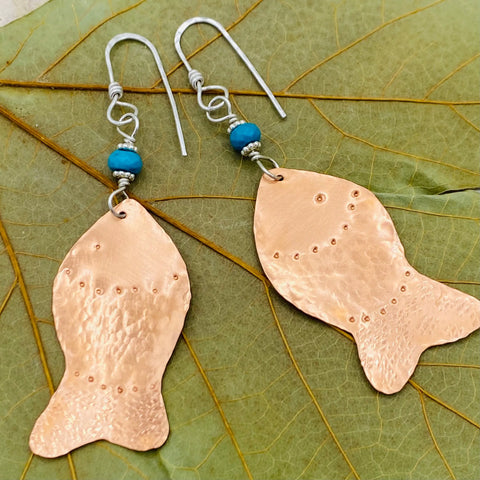 The JANECKA Copper Hand Forged Fish With Turquoise Earrings by Janecka feature hand forged copper, turquoise stones, wire hooks, and delicate dotted details, displayed on green leaves for a unique artisanal jewelry touch.
