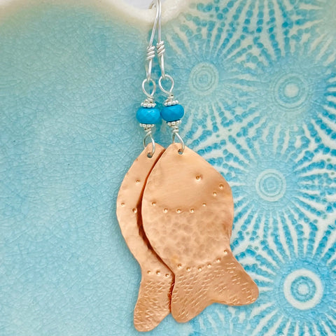 Janecka’s JANECKA Copper Hand Forged Fish With Turquoise Earrings feature textured copper fish and turquoise beads, shown on a blue patterned ceramic background—a striking piece of handmade jewelry.