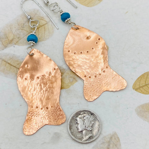 JANECKA Copper Hand Forged Fish With Turquoise Earrings by Janecka, shown beside a U.S. dime for size, feature unique fish tail shapes with turquoise accents—distinctive handmade jewelry on a leaf-patterned background.