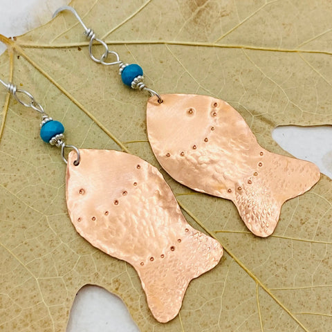 JANECKA Copper Hand Forged Fish With Turquoise Earrings by Janecka feature wire hooks, dotted fish detailing, and small turquoise beads. Displayed on a pale textured leaf, these unique handmade earrings are a standout jewelry piece.