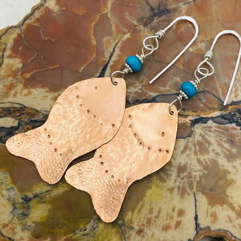 The JANECKA Copper Hand Forged Fish With Turquoise Earrings by Janecka feature textured copper fish, turquoise beads, and silver hooks—handmade jewelry displayed on a marbled stone background.