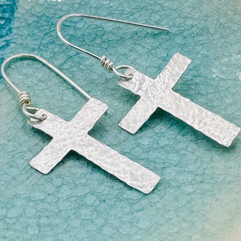JANECKA Sterling Silver Cross Earrings by Janecka—hand-forged with a hammered texture, shown on a light blue background. A unique anniversary gift or stylish sterling silver accessory.