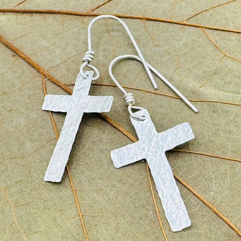 The Janecka Sterling Silver Cross Earrings are hand-forged and feature hooks, shown against a textured dried leaf backdrop—an elegant anniversary gift.