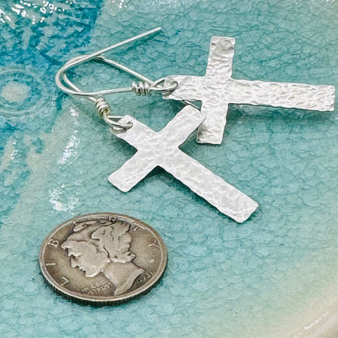 JANECKA Sterling Silver Cross Earrings, Hand Forged by Janecka, are shown beside a U.S. dime for size reference on a blue-green textured surface—making these elegant earrings an ideal anniversary gift.