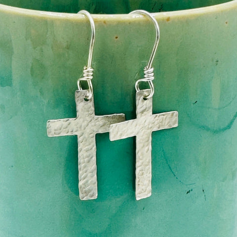 JANECKA Sterling Silver Cross Earrings, hand forged by Janecka, hang on wire hooks against a light green ceramic backdrop—an elegant anniversary gift.
