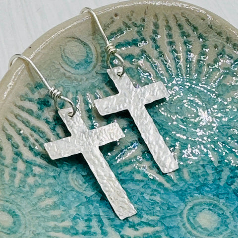 JANECKA Sterling Silver Cross Earrings, Hand Forged by Janecka, with hooks, are displayed on a blue and beige ceramic dish—an elegant anniversary gift idea.