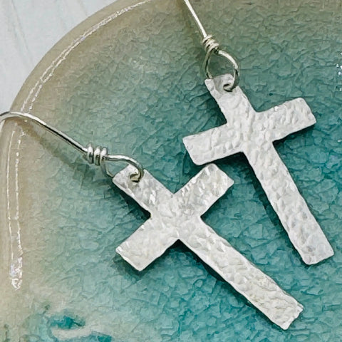 A close-up of JANECKA Sterling Silver Cross Earrings, Hand Forged by Janecka, featuring hook backs and displayed on a blue-green ceramic surface—an elegant anniversary gift idea.