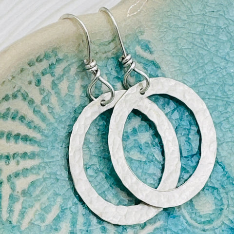JANECKA Sterling Silver Hand Forged Hoop Earrings by Janecka with a hammered finish and French hooks rest on a textured turquoise ceramic dish.