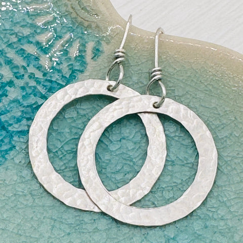 JANECKA Sterling Silver Hand Forged Hoop Earrings by Janecka, featuring artisan craftsmanship with ear wires, are displayed on a textured blue-green ceramic surface.