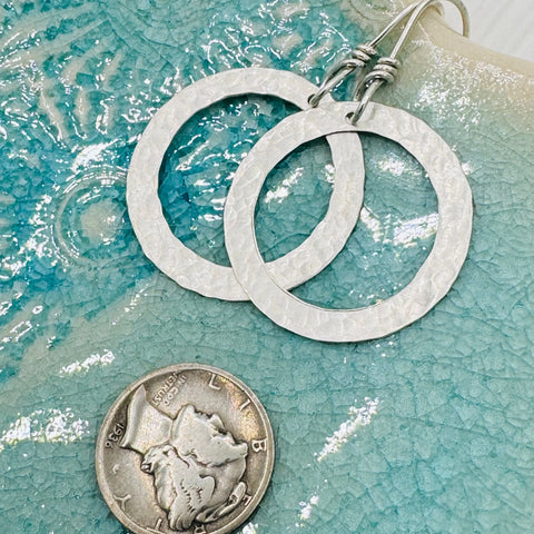 JANECKA Sterling Silver Hand Forged Hoop Earrings by Janecka, artisan crafted jewelry, shown beside a U.S. dime for scale on a textured blue and white ceramic surface.