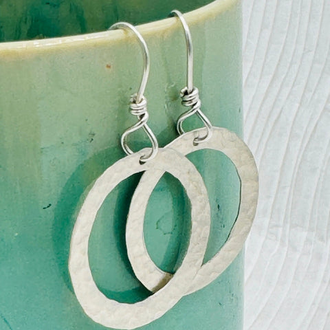 JANECKA Sterling Silver Hand Forged Hoop Earrings by Janecka, featuring elegant hook clasps, hang from a green and cream ceramic cup—an exquisite display of artisan jewelry craftsmanship.