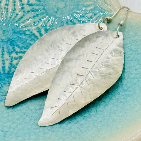 The JANECKA Sterling Silver Hand Forged Leaf Earrings by Janecka feature hammered texture and etched veins, shown on a turquoise ceramic dish with a textured pattern.