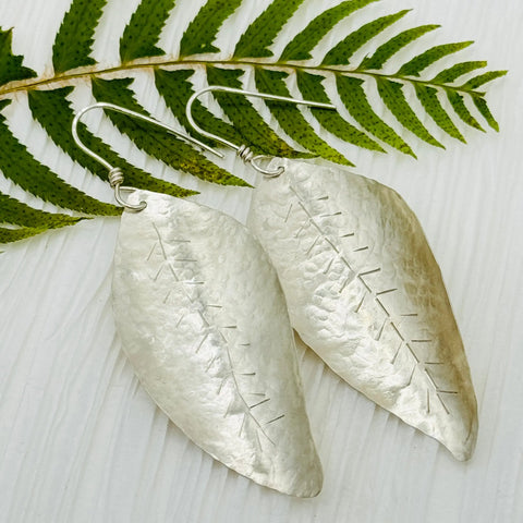 JANECKA Sterling Silver Hand Forged Leaf Earrings by Janecka, featuring an elongated leaf shape with hammered texture and etched vein details, are displayed on a white surface with a green fern leaf above them.